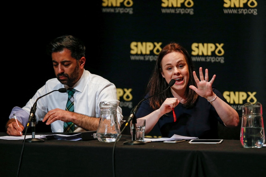 Forbes, right, gestures as she speaks into a microphone fitted to a long table. Yousaf sits beside her