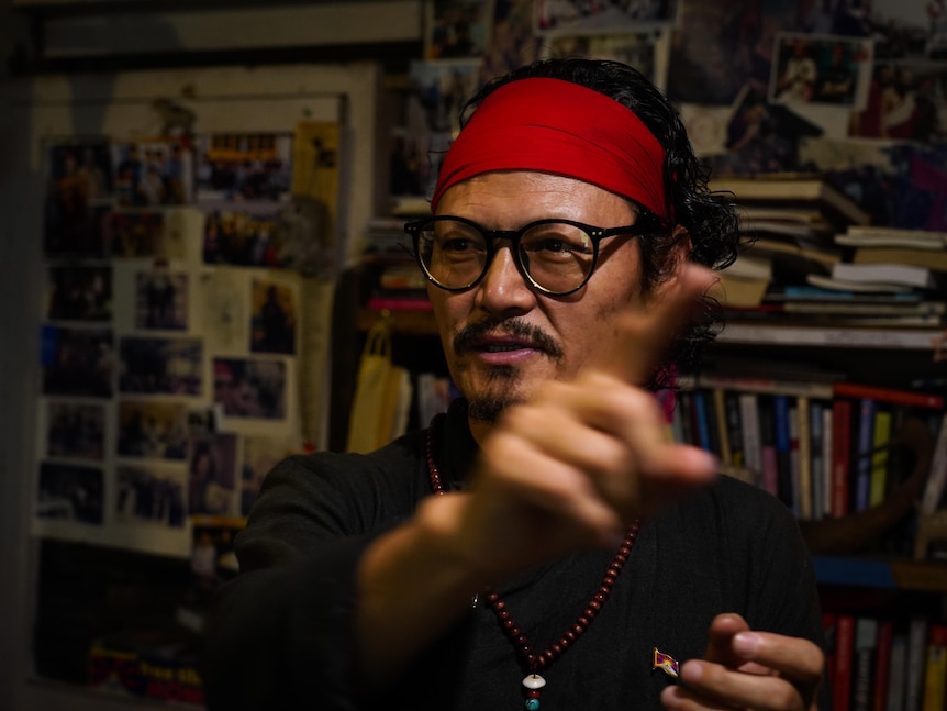 A man in a black shirt and red bandana gestures as he speaks in front of a messy bookshelf