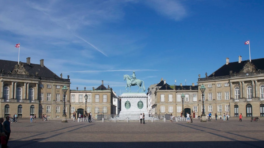 Wide shot of large palace buildings surrounding a square with a statue in the centre.