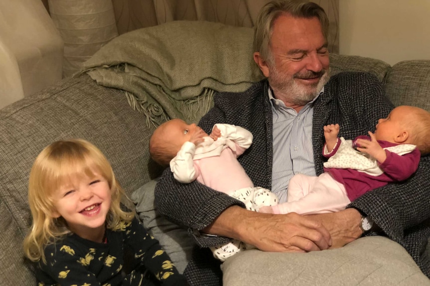 A man with a beard is smiling big holding twin baby girls in his arm as he sits on a couch. A toddler is smiling in foreground
