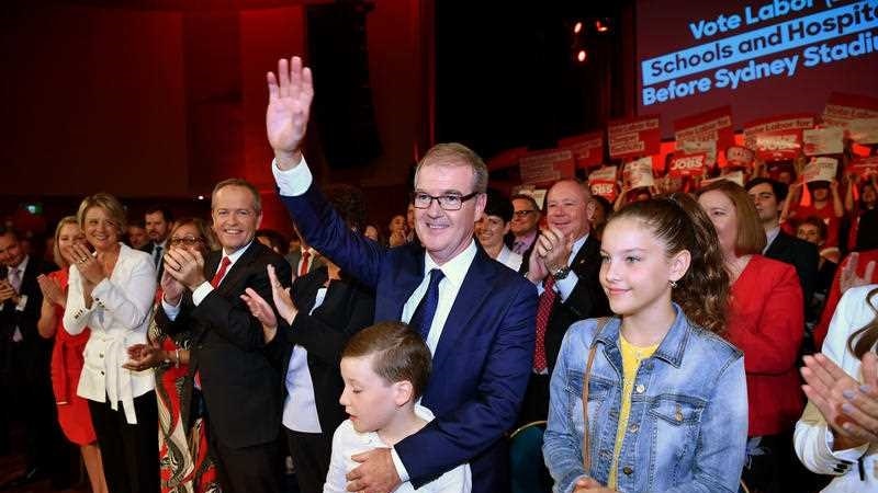 A man in a suit waves, while smiling adults and children stand closely around him.