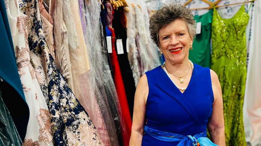 Lady wearing blue dress stands in her clothes shop in front of a number of her dresses on the rack