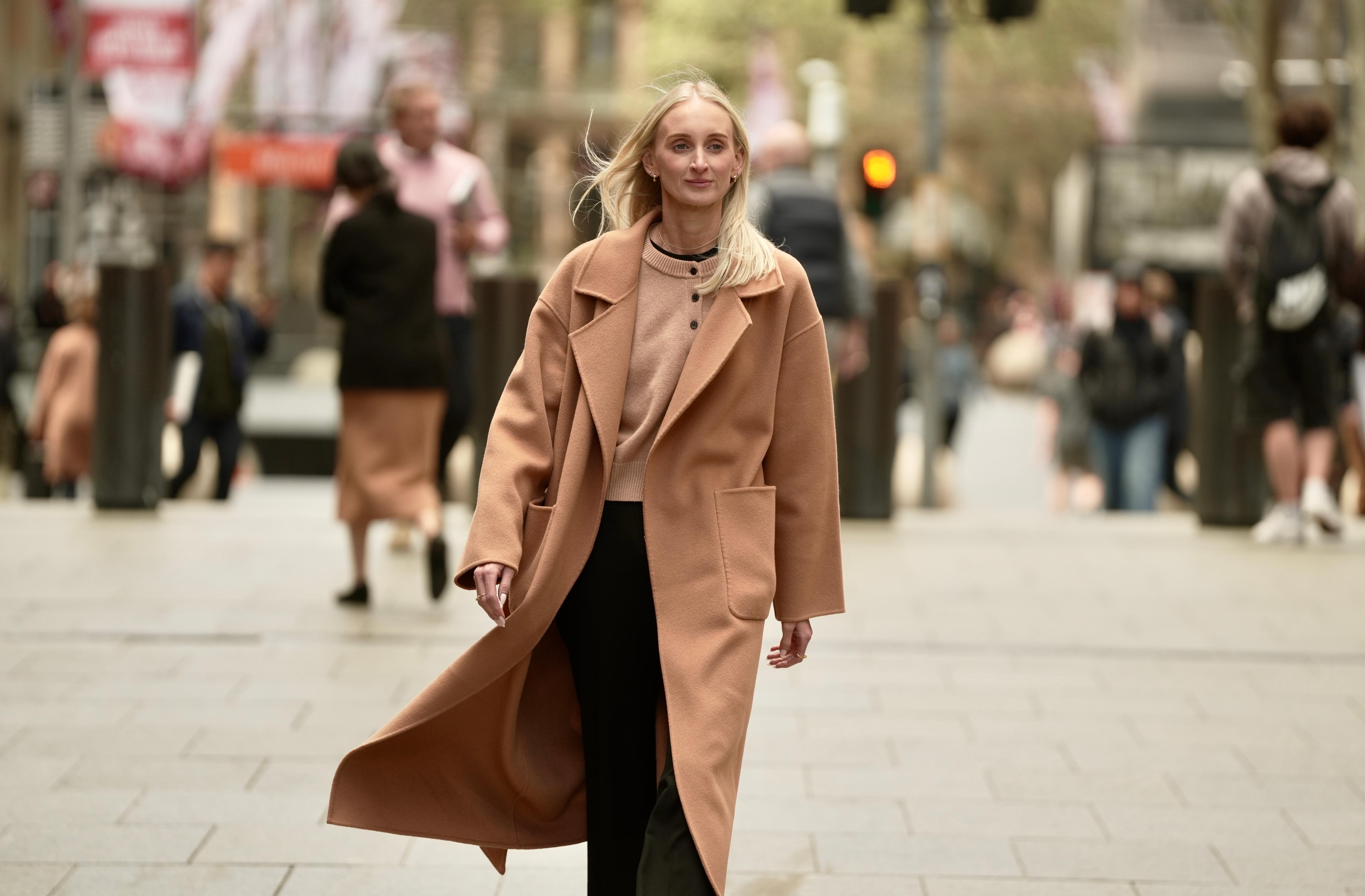 Samantha Bouffler, who has blonde hair and blue eyes, walks through a busy CBD plaza wearing a tan coat and black pants
