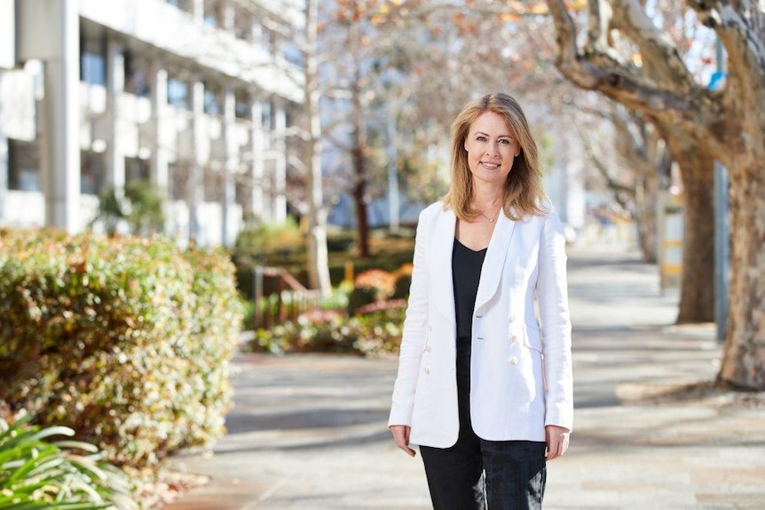 A wide shot of Di standing under deciduous trees wearing a white jacket and black shirt.