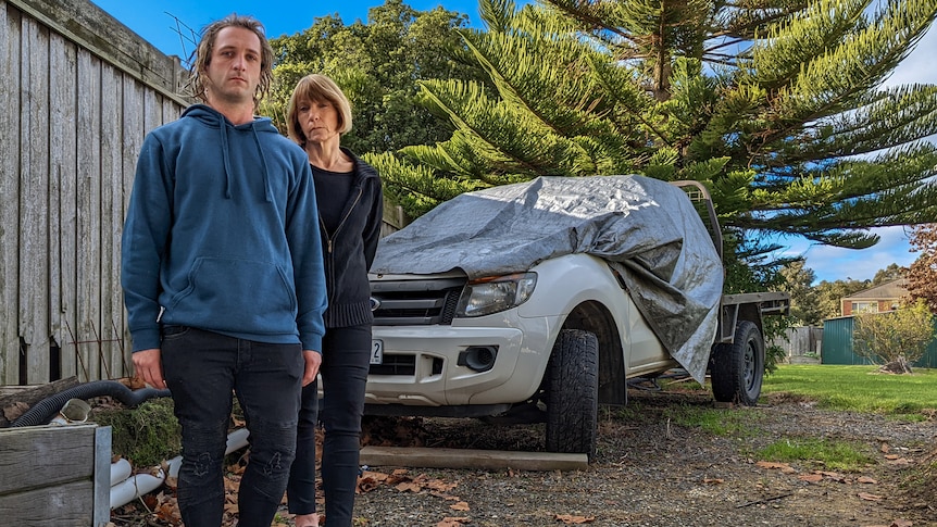 A man in his twenties and his mum stand in front of a white ute that has been covered in a tarp to protect if from the weather.