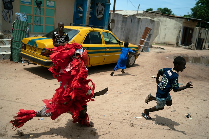 a man dressed in a red Kankurang suit chases two boys while a man leans on a yellow car in the background