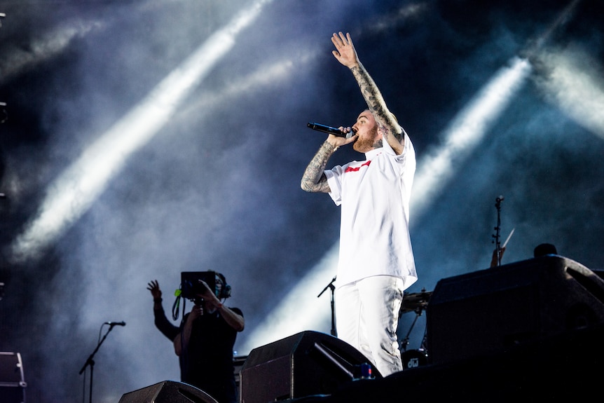rapper mac miller holds a microphone on a stage with lights in the background. he has one hand up and is dressed in all white