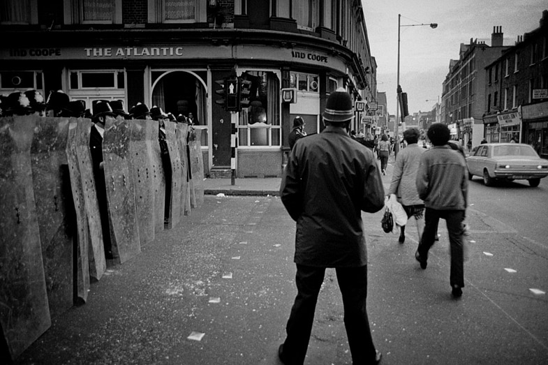 POliceman stands near a pub called The Atlantic with a row of riot police behind shields next to him people walking on road