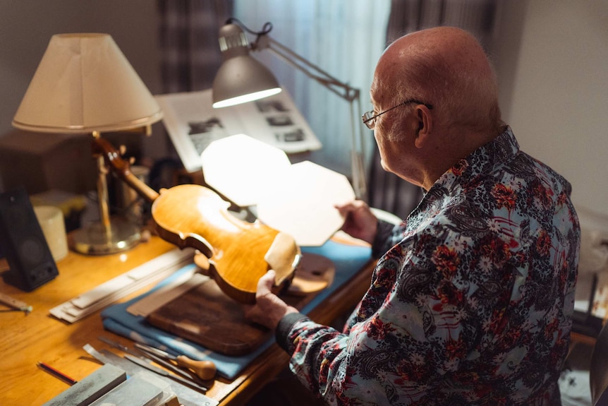 A bald man wearing a dark floral shirt sits down at a desk tuning up a violin
