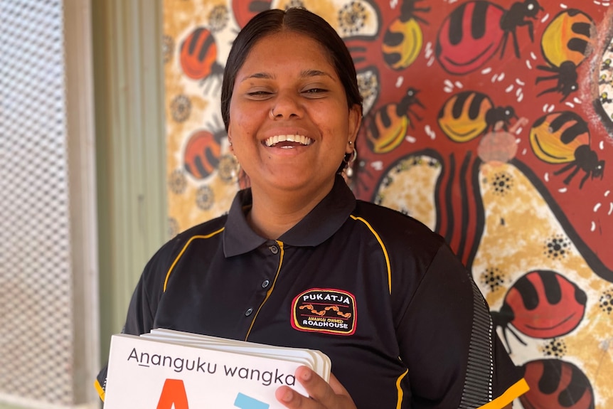 A young Aboriginal woman smiling as she holds a brightly coloured picture book.