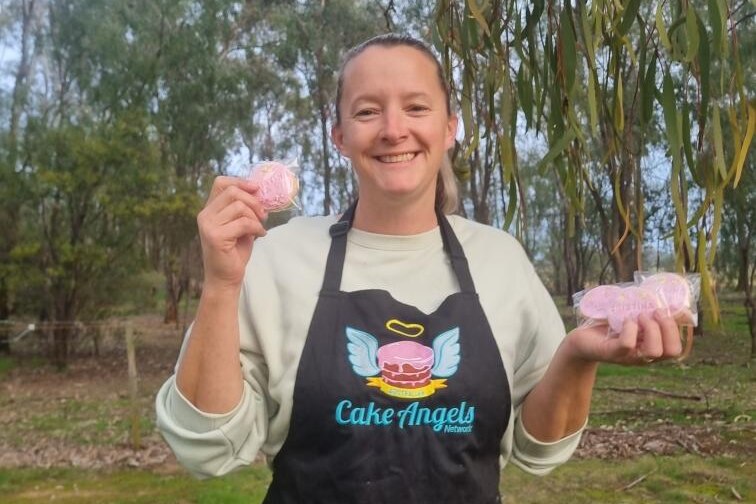 A woman stands outside surrounded by trees in a black apron reading 'Cake Angels'.