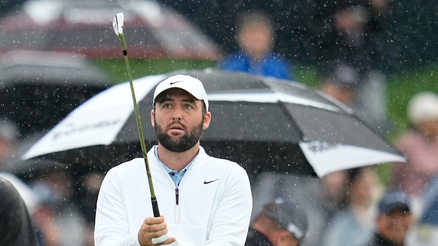 Scottie Scheffler holds his golf club while standing in the rain on the practise range.