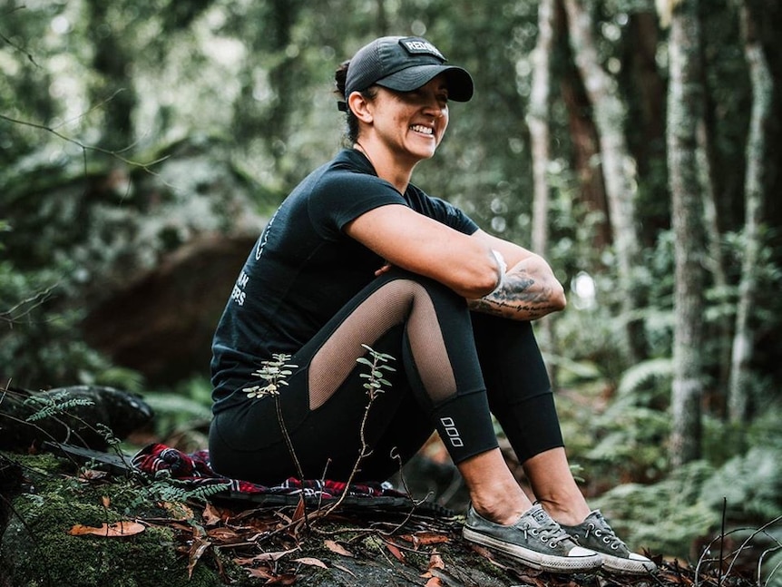 A woman wearing exercise clothing sits on a rock in a forest.