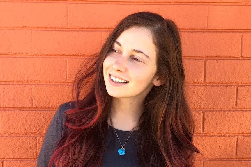 A smiling young woman with long brown hair, tinged with red, stands in front of an orange wall.