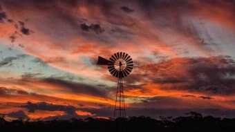 A windmill in the outback in front of clouds lit up red and orange by a sunset.