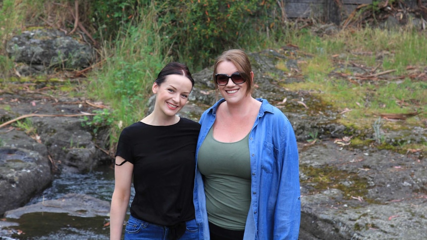 Two women smiling, one wearing sunglasses, standing by a rivulet