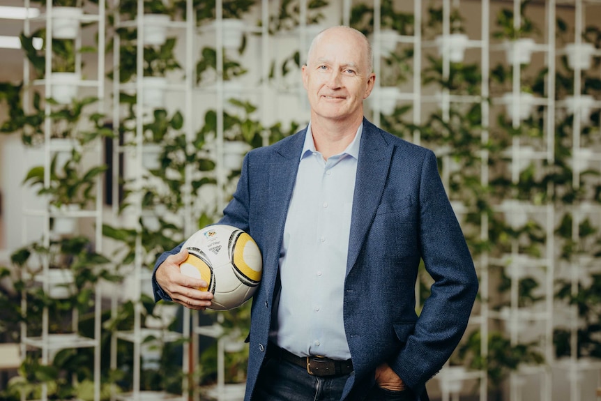 A man in a suit jacket holds a soccer ball in front of a wall of plants inside an office.