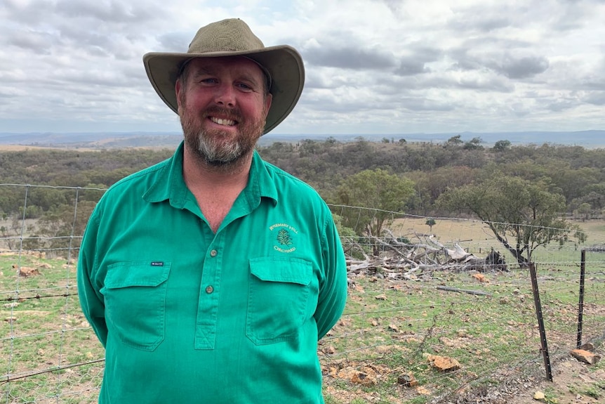 A man in a green shirt and a hat stands in front of a fence and paddocks.