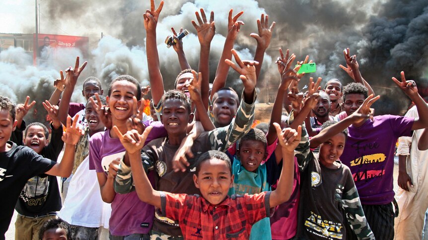 A group of young, smiling African boys in colourful western clothing raise victory signs as smoke billows behind in street