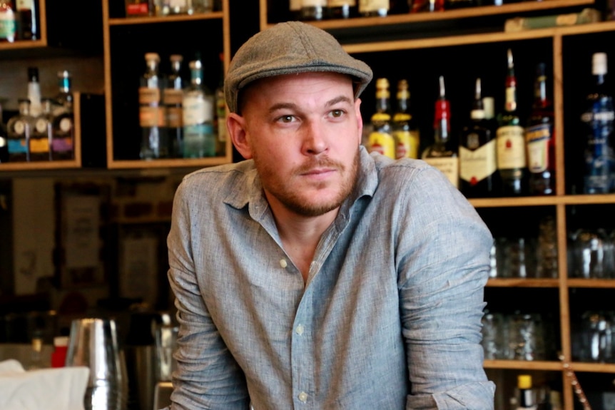 A mean wearing a cap and open-necked shirt stands leaning on a bar with bottles of alcohol behind him.