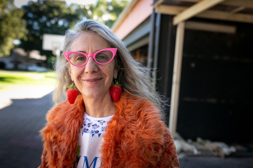 A woman wearing pink glasses, strawberry drop earrings and a furry orange jacket smiles.