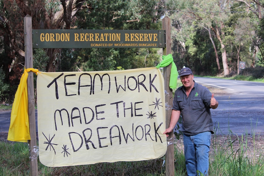 A man has his thumbs up next to a sign that reads, "Teamwork made the dreamwork".