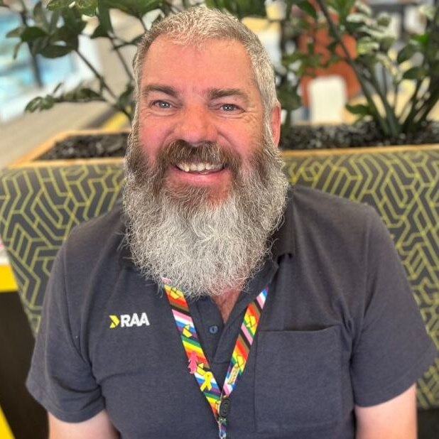 man with short grey hair and long grey beard smiling on a couch in black polo shirt and colourful lanyard