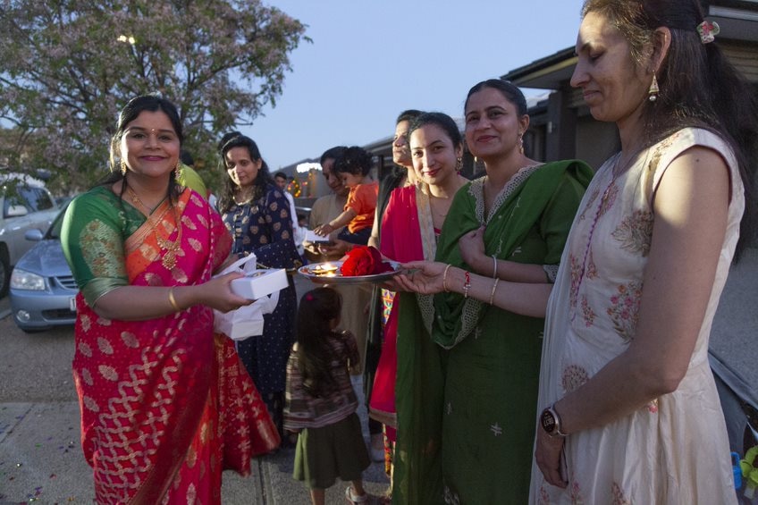 A woman in a bright saree hands out a box to a group of women in Indian traditional clothes.