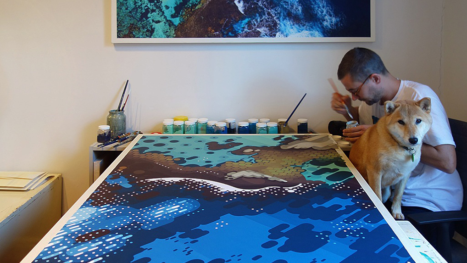 Artist Amok Island paints an artwork on a large table with a dog sitting in his lap.