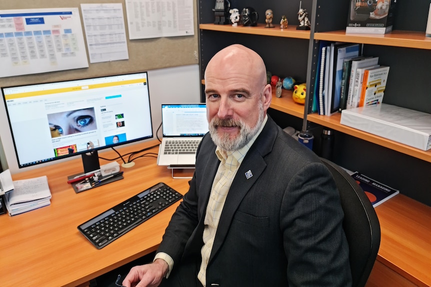 A middle aged man in a suit sits in front of a computer at a desk.