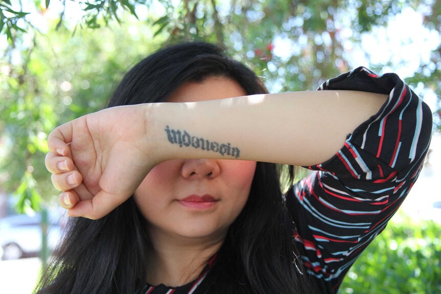 A woman holds up her arm, tattooed with the word "Indonesia", in front of her eyes against a leafy background.