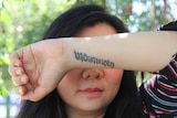 A woman holds up her arm, tattooed with the word "Indonesia", in front of her eyes against a leafy background.