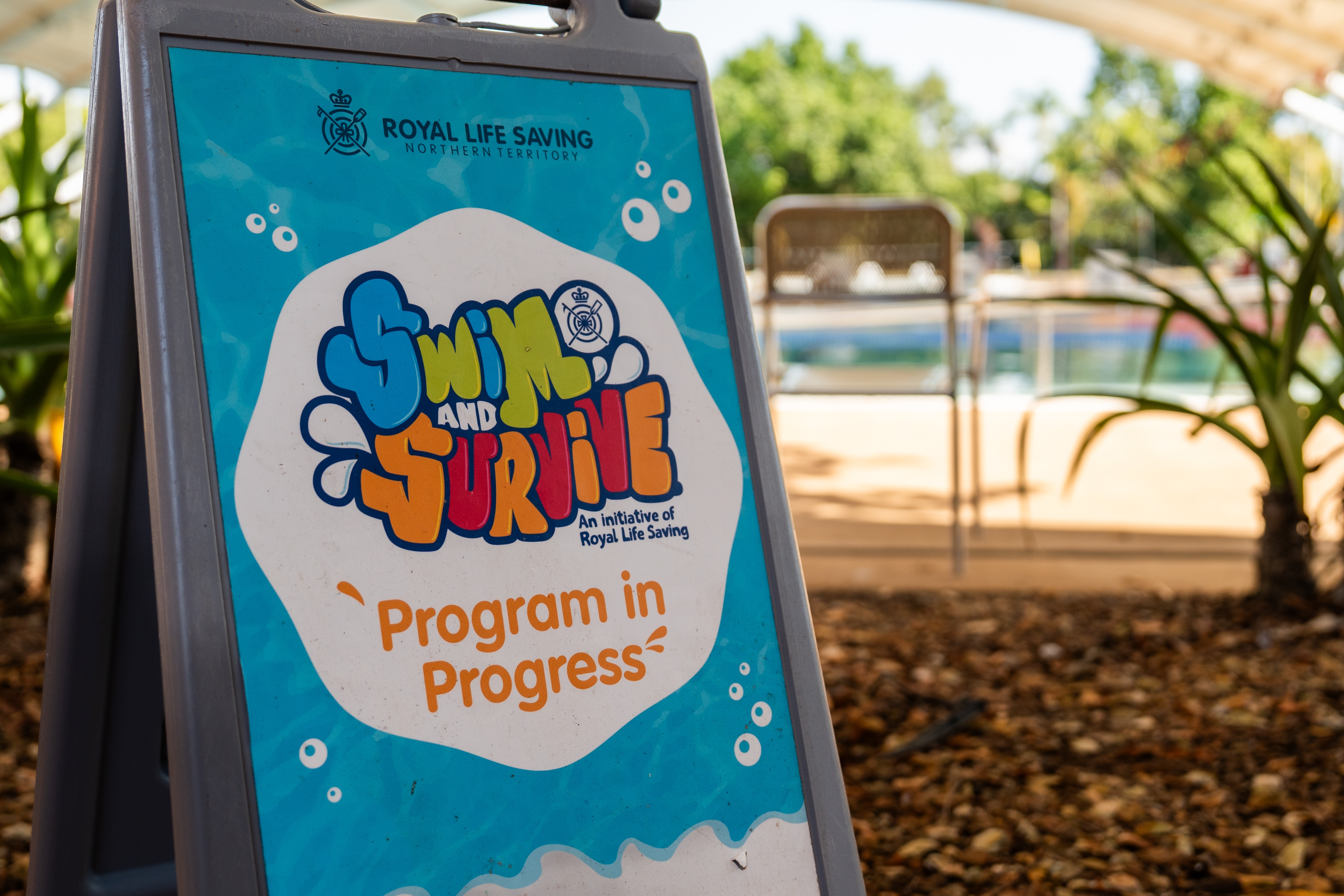 A sign at the entrance of a community pool reads: Royal Life Saving, swim to survive, program in progress