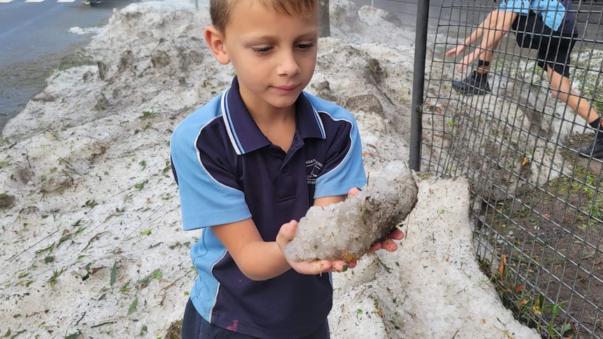Young boy in school uniform holds chunk of hail bigger than his two hands. 