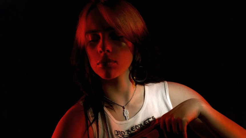 Image of Billie Eilish; Billie wear a white singlet with 'No Regrets' on it, is photographed against black backdrop
