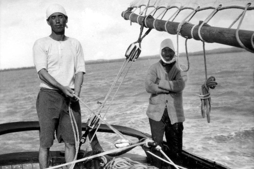 A black and white photo of two men standing on a ship, one is holding a rope connected to the tiller.