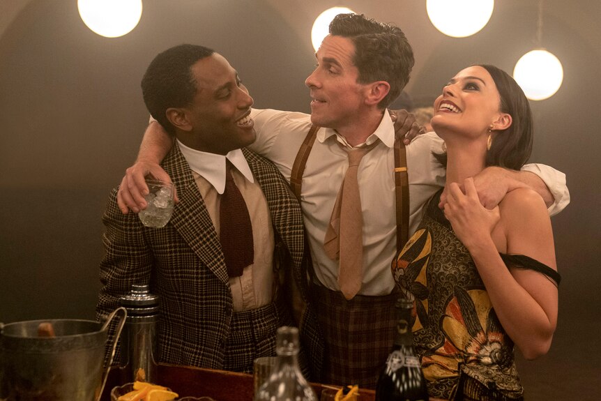 Three people embracing side by side, during a party in dapper 1930's outfits, a Black man, white man and white woman