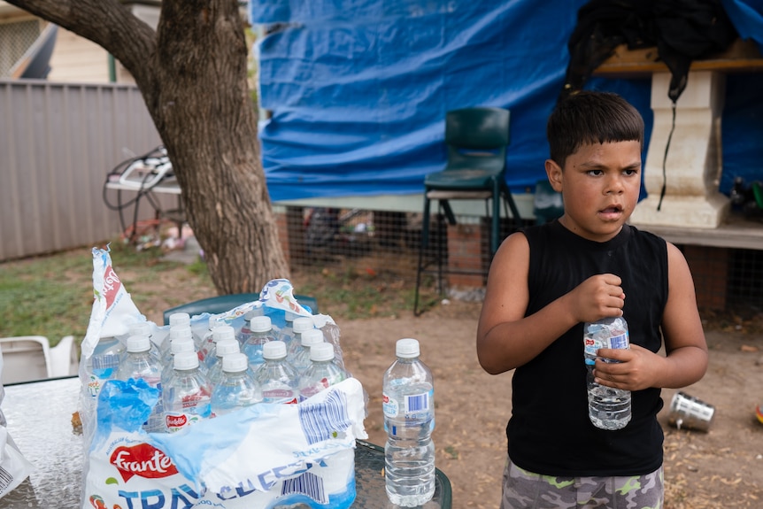 Young Indigenous boy in a black singlet holds a water bottle, next to a table with a pallet of water bottles.