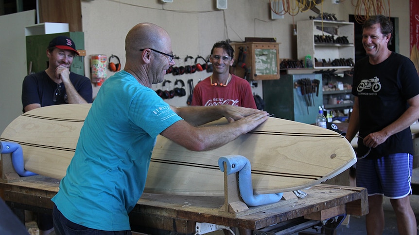 Three men standing around a woodworking teacher sanding the edges of a wooden surfboard in a workshop.