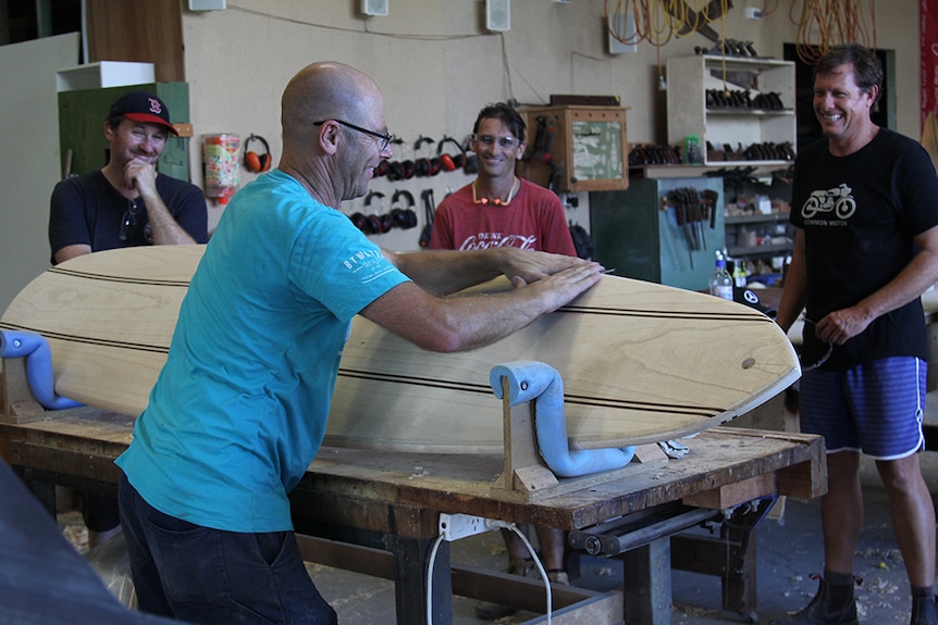 Three men standing around a woodworking teacher sanding the edges of a wooden surfboard in a workshop.