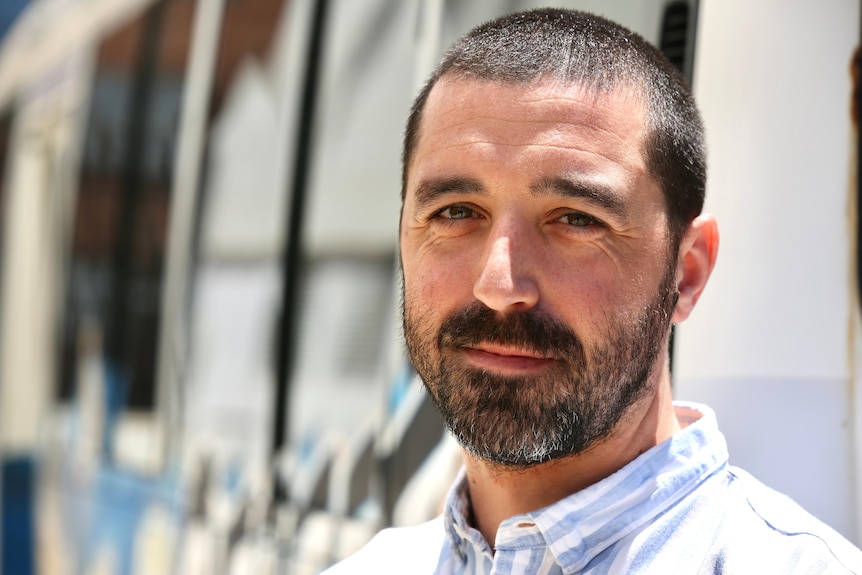 A man with short dark hair and beard looking into the camera standing before a bus emblazoned with street art.