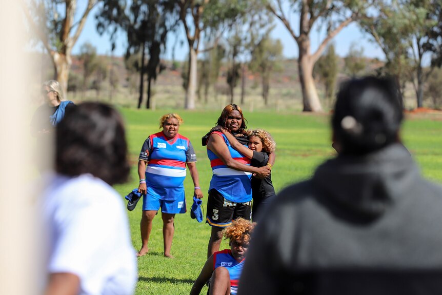 Two Aboriginal women hug while watching an address on a football ground from other Aboriginal women