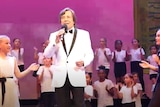 John Paul Young in a white suit on stage with school students performing around him  
