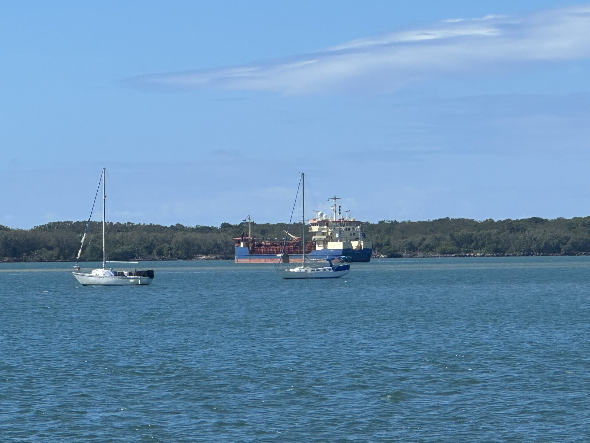 A large barge and two sailing boats anchored in the ocean near a bushy coastline.