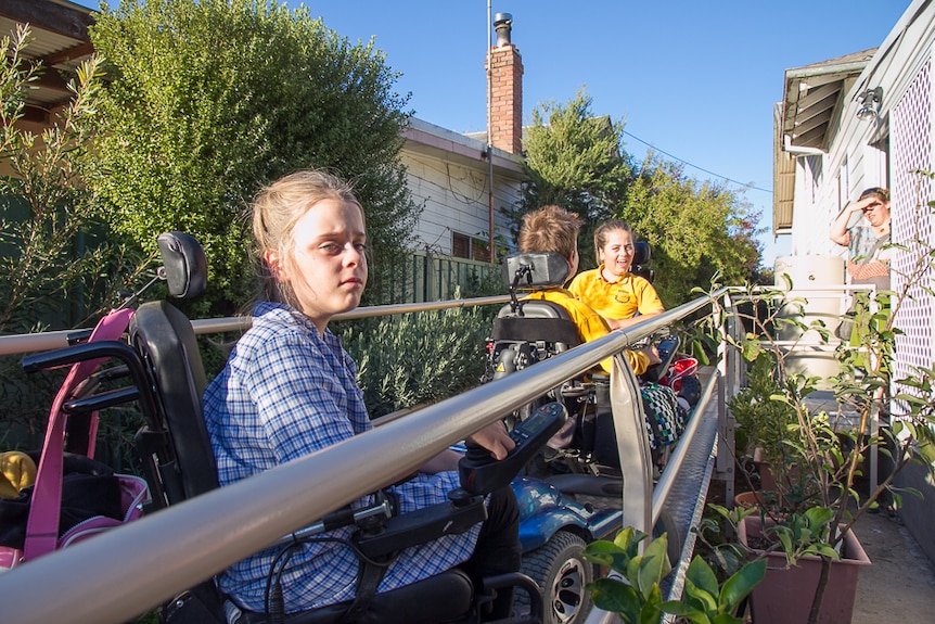 Three children in school uniforms and in power wheelchairs on a ramp leading to a house