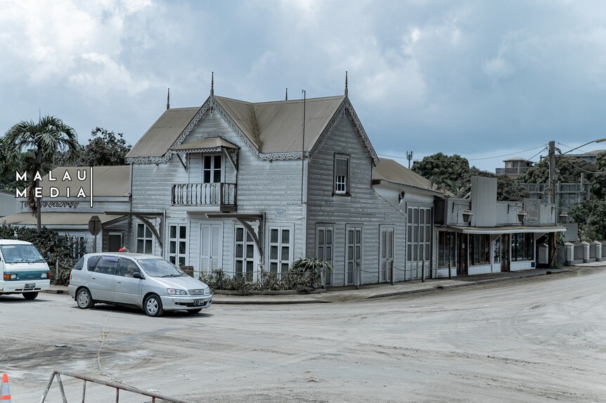 White colonial house on corner of road intersection, car sits outfront to turn left. Sky is grey. Volcanic ash in air. 