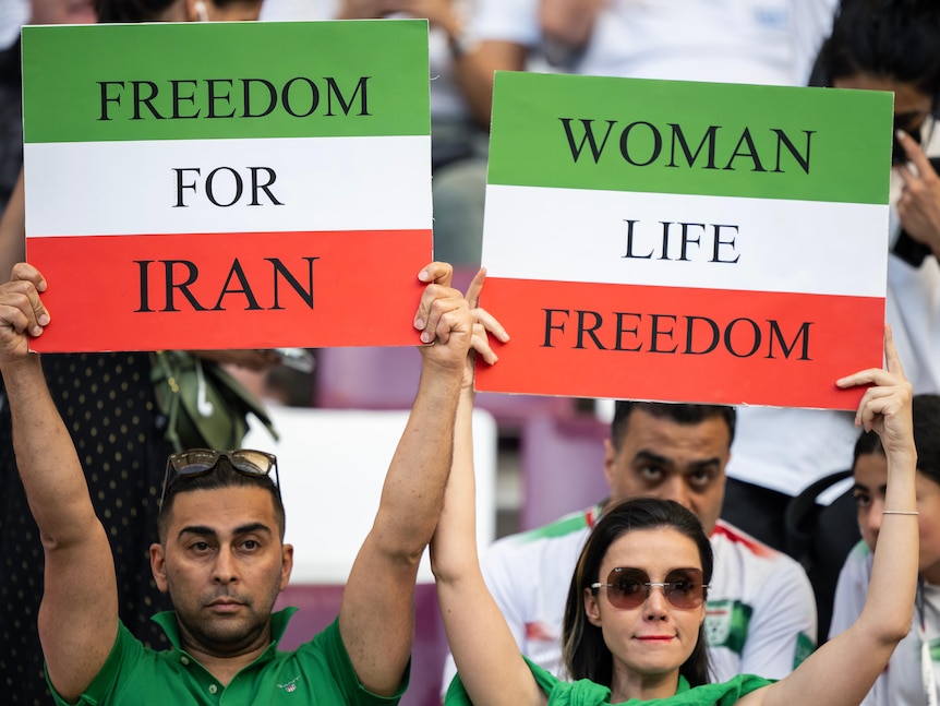 A man and a woman standing in a crowd hold up signs saying 'freedom for Iran' and 'woman life freedom'. 