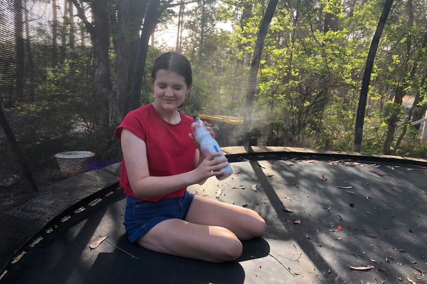 A teenage girl sits on a trampoline and sprays herself with a water spray bottle.