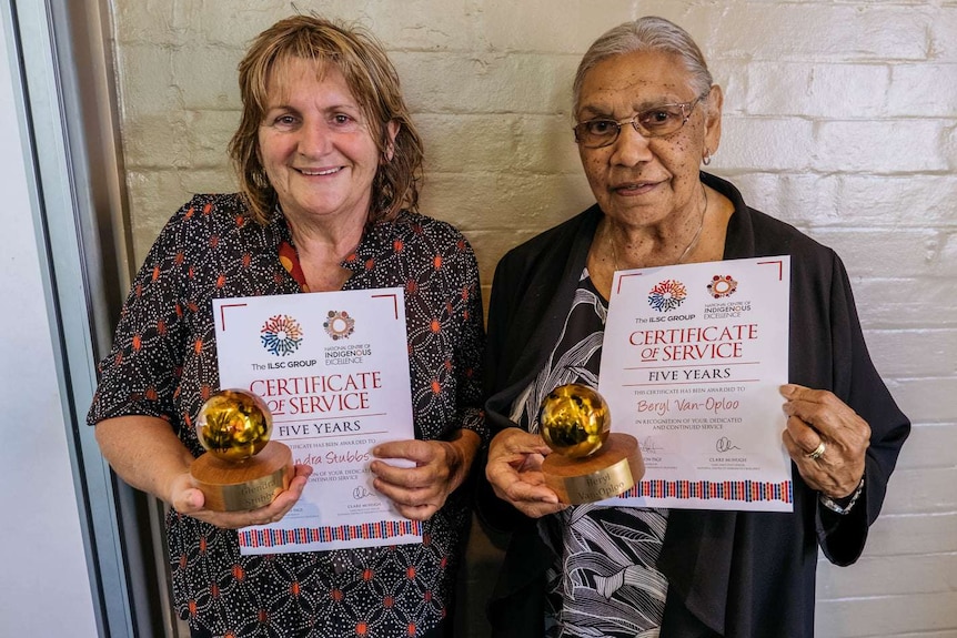 Two Indigenous women holding certificates and trophies look towards the camera.