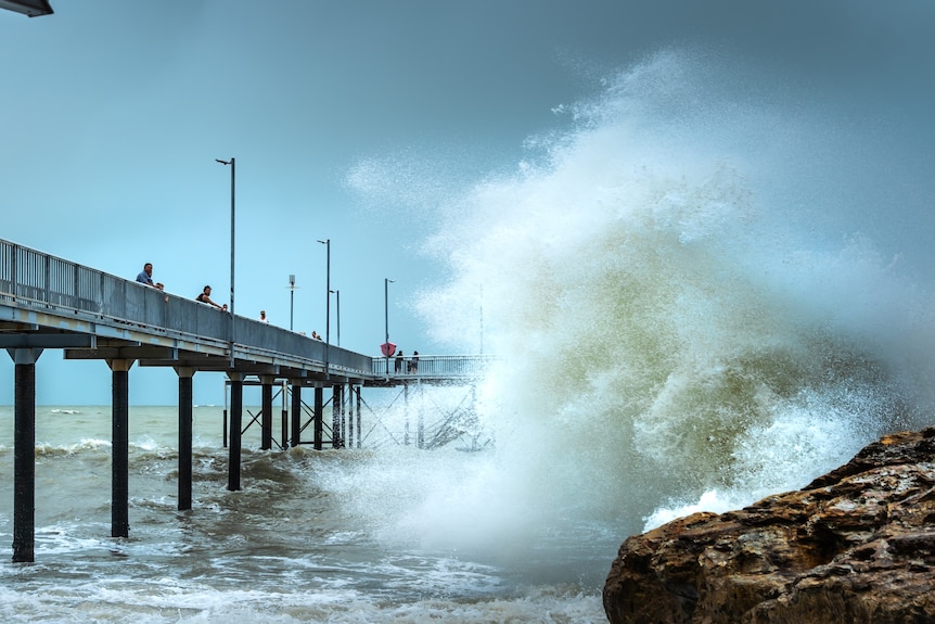 A large wave washes over a jetty as dark clouds loom in the background.